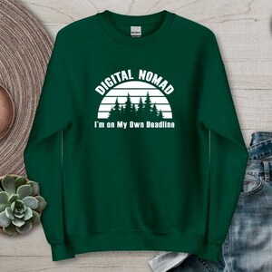 Może przedstawiać: Leśna zielona bluza z napisem "DIGITAL NOMAD" nad grafiką lasu i frazą "I'm on My Own Deadline". Bluza jest pokazana na neutralnej powierzchni z zegarkiem i rośliną sukulentową.