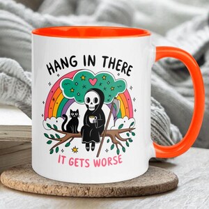 Pu&ograve; includere: Una tazza da caff&egrave; in ceramica bianca con un bordo arancione. La tazza presenta un'illustrazione in bianco e nero di uno scheletro che tiene una tazza di caff&egrave;, seduto su un ramo con un gatto nero. Il testo sulla tazza dice "Hang in there, it gets worse".