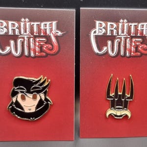 Peut inclure: Deux épingles en émail sur des cartes rouges. L'une représente un personnage de dessin animé aux cheveux noirs et un col noir. L'autre est un trident noir et doré. Le texte "BRÜTAL CUTIES" est en haut de chaque carte.