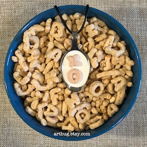 May include: A silver spoon necklace with a white and pink frosted donut and a white and pink frosted letter X inside the spoon. The spoon is surrounded by a bowl of alphabet cereal.