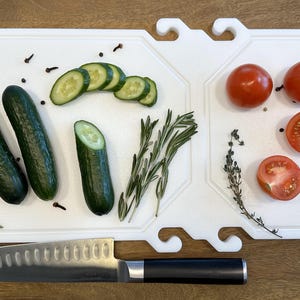 Könnte beinhalten: Ein weißes Schneidebrett mit einer Vielzahl frischer Zutaten. Gurken, geschnittene Gurken, Tomaten und Kräuter sind auf dem Brett angeordnet. Ein Edelstahlmesser mit schwarzem Griff ist ebenfalls vorhanden.