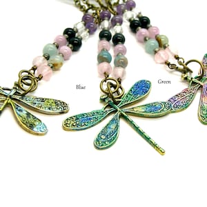 May include: Three dragonfly necklaces with beaded strands. The dragonfly pendants are in black, blue, and green, with intricate wing designs. The beads are in shades of pink, purple, and green. The word "Black", "Blue", and "Green" are written above each dragonfly.
