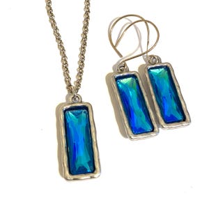 May include: A jewelry set consisting of a necklace and a pair of drop earrings. Each piece features a rectangular, faceted blue crystal set in a textured, silver-toned metal frame. The necklace includes a silver-toned chain, while the earrings have hook-style backings.