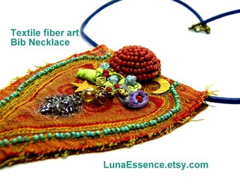 May include: A textile fibre art bib necklace featuring a rust-coloured fabric base with frayed edges, adorned with turquoise beads, a red beaded dome, a gold star, a gold moon, and various fabric accents. Text reads: Textile fibre art Bib Necklace.