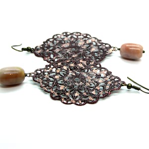 May include: A pair of ornate earrings with openwork filigree designs in a dark reddish-brown colour. Each earring has a light peach-coloured, oblong bead. The earrings have a vintage, bohemian aesthetic.