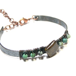May include: A handcrafted bracelet with a distressed metal band. The bracelet features a square, brown stone and small, turquoise beads. The beads are wire-wrapped, adding a unique touch to this piece of jewellery.