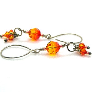 May include: A pair of dangle earrings featuring faceted orange and yellow beads. The earrings have a silver-tone metal hook and are accented with smaller orange and pink beads. The design is set against a white background.