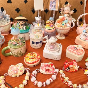 May include: A collection of decorative figurines and accessories, including mugs, music boxes, and necklaces. The figurines feature detailed designs and pastel colors. The items are arranged on an orange surface, creating a whimsical display.