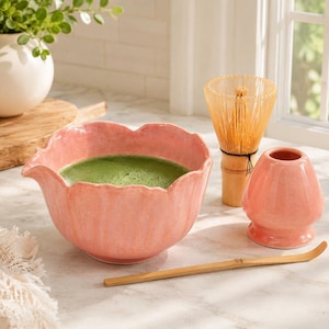May include: A pink ceramic matcha bowl with a scalloped rim containing green tea, accompanied by a light brown bamboo whisk, a matching pink ceramic whisk holder, and a long, thin, curved bamboo tea scoop for preparing matcha tea.