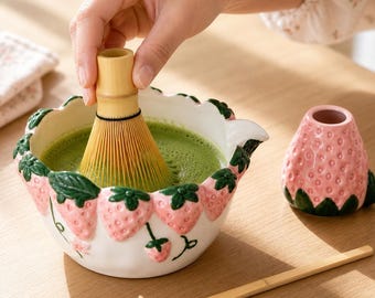 May include: A white ceramic matcha bowl featuring a raised strawberry pattern with green leaves, containing green tea. A bamboo whisk is inside the bowl, accompanied by a matching pink strawberry-shaped whisk holder and a bamboo tea scoop.