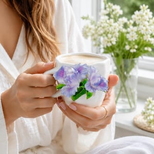 Puede incluir: Una taza de cerámica blanca con un diseño floral tridimensional en el exterior. La taza está decorada con flores esculpidas de color morado y azul con hojas verdes adheridas al costado. El interior de la taza contiene una bebida color café claro.