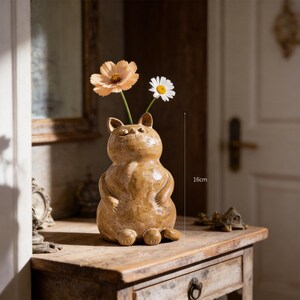 Può includere: Un vaso in ceramica color marrone chiaro a forma di gatto con finitura maculata, dotato di due piccole orecchie e un corpo arrotondato. Il vaso contiene due fiori, uno color pesca e uno bianco con centro giallo. Misura 16 cm in altezza e 9 cm in larghezza.