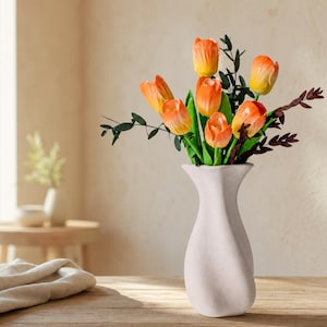 May include: A bouquet of orange and yellow tulips with green leaves and dark stems arranged in a tall, off-white, hourglass-shaped ceramic vase with a smooth, matte finish. The flowers feature vibrant petals with subtle color gradients.