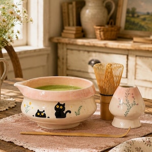 May include: A ceramic matcha bowl with a pink rim and black cat illustrations, accompanied by a bamboo whisk, a whisk holder with floral patterns, and a bamboo scoop. The bowl contains green tea and features small yellow flower accents.