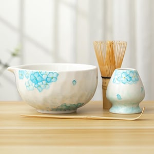 Può includere: Un set in ceramica bianca per la cerimonia del tè matcha, composto da una ciotola, un porta frusta e una frusta in bambù. La ciotola e il supporto sono decorati con motivi floreali blu. Un cucchiaio di bambù è posto davanti al set.