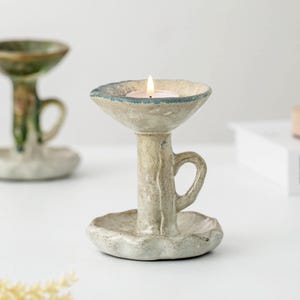 Può includere: Un portacandele in ceramica fatto a mano con una candela accesa. Il supporto è beige con accenti blu e un piccolo manico. La base ha un design smerlato. Un altro supporto simile è sullo sfondo.