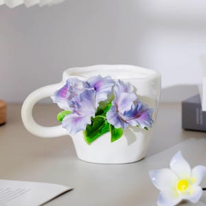 May include: White ceramic mug with a sculpted floral design featuring purple and blue irises and green leaves. The mug has a curved handle and sits on a light-colored surface.