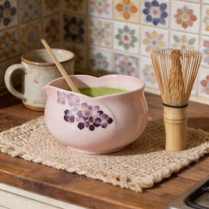 Könnte beinhalten: Eine hellrosa Keramikschale mit einem violetten Hortensien-Blumenmuster, die grünen Matcha-Tee enthält. Ein Bambusbesen steht rechts daneben und ein heller Bambuslöffel liegt in der Schale. Ein gesprenkelter Keramikbecher steht im Hintergrund.