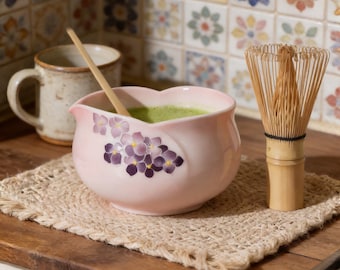May include: A light pink ceramic bowl with a purple hydrangea floral pattern, containing green matcha tea. A bamboo whisk stands to the right, and a light-coloured bamboo spoon rests inside the bowl. A speckled ceramic mug sits in the background.