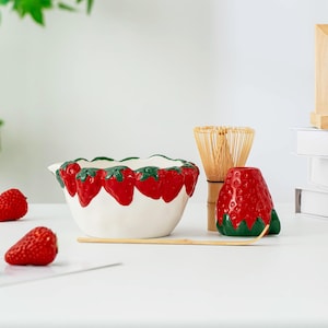 Puede incluir: Un cuenco de cerámica blanco con un diseño de fresas alrededor del borde, un recipiente para té con forma de fresa roja, un batidor de bambú y una cuchara de madera. El cuenco y el recipiente tienen detalles verdes. Fresas frescas esparcidas.