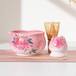 Op de afbeelding: Een roze keramische matcha kom met een bloemenontwerp, een bamboe klopper en een kleine roze keramische container met een bloemenontwerp. De kom heeft een tuit en is versierd met roze kersenbloesems en groene bladeren.