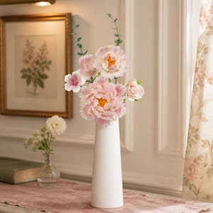 Peut inclure: Un grand vase cylindrique blanc et lisse contient un bouquet de fleurs artificielles, comprenant de grandes pivoines roses aux pétales froncés et cœurs jaunes, des fleurs roses plus petites et des tiges d'eucalyptus aux feuilles vertes.