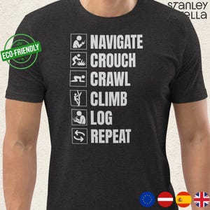 Op de afbeelding: Donkergrijs t-shirt met witte tekst en iconen. De tekst luidt "NAVIGATE, CROUCH, CRAWL, CLIMB, LOG, REPEAT." Een "ECO-FRIENDLY" sticker staat op de linkerschouder. Het shirt is van Stanley Stella.
