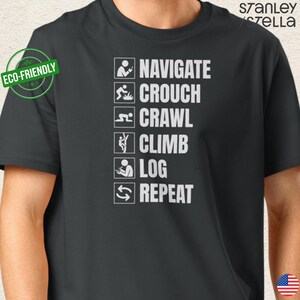 Peut inclure: T-shirt noir avec texte et icônes blancs. Le texte indique "NAVIGATE, CROUCH, CRAWL, CLIMB, LOG, REPEAT". Un autocollant "Eco-Friendly" est sur le côté gauche. La marque "stanley stella" est en haut à droite.