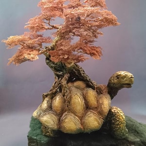 May include: A sculpture of a tortoise with a textured, golden-brown shell supporting a copper-colored wire tree. The tortoise has a green-toned head and legs, and it stands on a dark, mossy-textured base that rests on a circular platform.