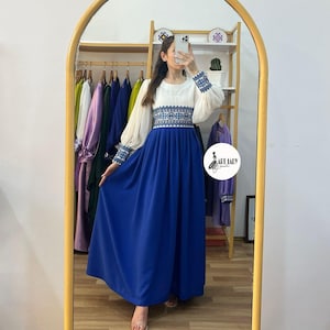 May include: A white and blue embroidered dress with a blue skirt and puffy white sleeves. The dress has a fitted bodice and a flowing skirt. The model is wearing the dress in a shop setting.