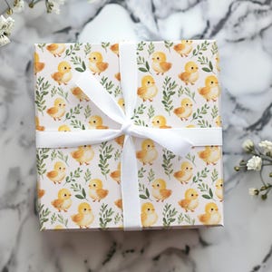 May include: A wrapped gift box featuring a repeating pattern of yellow chicks and green leaves on a white background. The box is tied with a white ribbon. The overall theme is spring or Easter.