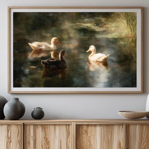 May include: A framed art print depicts three ducks floating on a pond. Two white ducks with orange beaks and one brown duck are in the water. The artwork has a soft, painterly style with muted greens and browns, evoking a serene outdoor scene.