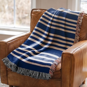 May include: A blue and white striped throw blanket with fringe detail draped over a brown leather armchair. The blanket features thick horizontal stripes in navy blue and cream. The fringe is a mix of black, red, and cream.