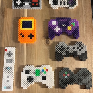 Peut inclure: Une collection de huit contrôleurs et consoles de jeux vidéo en pixel art réalisés avec des perles Perler colorées. Les contrôleurs comprennent un Nintendo Entertainment System, une Super Nintendo, une GameCube, une Xbox, une Playstation et une Wii. Les consoles comprennent une Game Boy et une télécommande Wii.