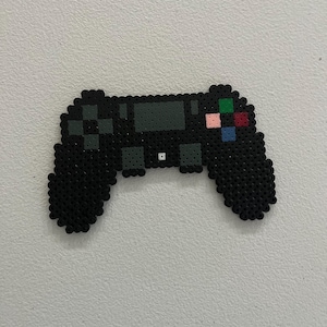 May include: Black pixel art video game controller with colorful buttons. The controller has a symmetrical design with two handles and various buttons, including a directional pad and action buttons. The controller is made of small, square pixels.