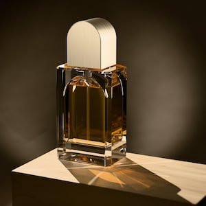May include: A rectangular glass perfume bottle filled with amber-colored liquid, featuring a thick clear base and a smooth, arched metallic gold cap. The bottle has a distinct internal arch design and reflects light onto the surface below.