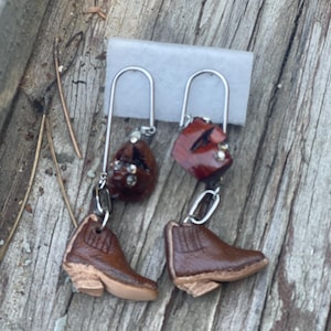 May include: A pair of dangle earrings featuring brown leather boot charms and red gemstone accents.