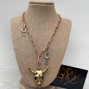 May include: A gold bull skull pendant necklace with a rose gold chain and silver charms. The charms include a horseshoe, a four-leaf clover, and a star. The necklace is displayed on a brown mannequin.