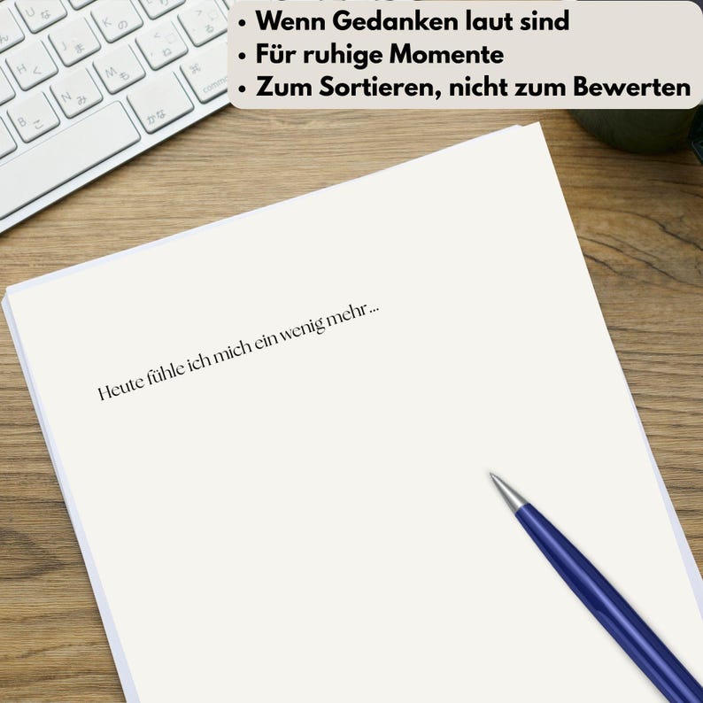 Op de afbeelding: Een wit vel papier met de tekst "Heute f&uuml;hle ich mich ein wenig mehr..." ligt op een houten bureau. Een blauwe pen ligt op het papier. De bovenkant van een toetsenbord is zichtbaar op de achtergrond, evenals de tekst "Wenn Gedanken laut sind", "F&uuml;r ruhige Momente" en "Zum Sortieren, nicht zum Bewerten".
