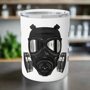 Puede incluir: Taza de viaje blanca con un diseño de máscara de gas negra. La máscara de gas tiene lentes grandes y una estética industrial detallada. La taza incluye el texto "S L O W B U R N" en formato vertical en el lado derecho.