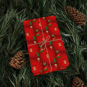 Pode incluir: Um presente embrulhado com papel vermelho com um padrão de renas, árvores de Natal e elfos. O presente é amarrado com barbante e repousa sobre agulhas de pinheiro, com pinhas visíveis.