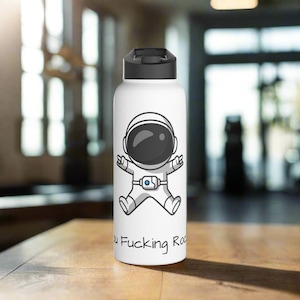 Könnte beinhalten: Weiße Wasserflasche mit schwarzem Deckel und einer Cartoon-Illustration eines Astronauten im Raumanzug. Der Text "You Fucking Rock" ist auf der Unterseite der Flasche aufgedruckt.