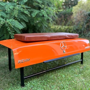 May include: Orange car trunk bench with a brown leather cushion and black metal legs. The trunk has a Mercedes-Benz logo and the word "REHM" printed on it.