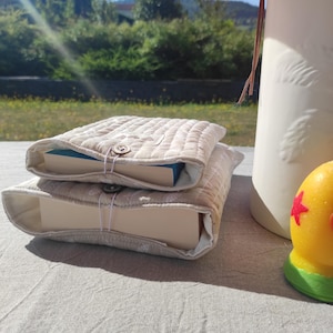 Peut inclure: Deux housses de livre en tissu matelassé beige avec un motif blanc subtil, chacune dotée d'une fermeture à bouton avec une boucle élastique. Les housses sont empilées, celle du haut contient un livre à tranche bleue et celle du bas un livre épais.