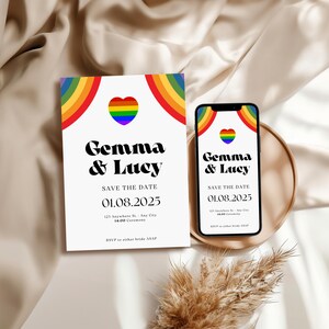 May include: A white invitation card and a smartphone display the text "Gemma & Lucy" with a rainbow heart design. The card and phone show the date 01.08.2025. The invitation is on a beige satin surface.