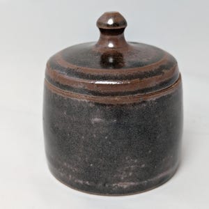May include: A small, round ceramic jar with a lid. The jar is dark gray with a speckled texture, and the lid is brown with a small knob on top. The lid has a darker brown band around the edge. The jar is likely used for storage.