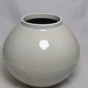 Puede incluir: Un jarrón grande, redondo, de cerámica blanquecina con una abertura ancha y un interior oscuro. El jarrón tiene un acabado liso y brillante y una superficie ligeramente texturizada. El jarrón es de color neutro y podría usarse con fines decorativos.