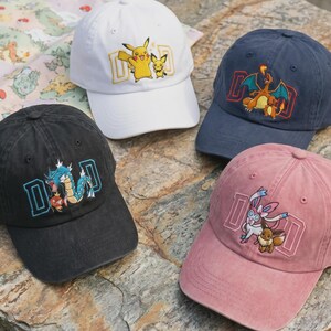 May include: Four baseball caps in white, navy blue, black, and pink. Each cap features a different Pokemon character design within a rectangular frame. The caps are displayed on a stone surface.