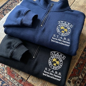 Könnte beinhalten: Zwei Sweatshirts mit Viertelreißverschluss, eines in Marineblau und eines in Schwarz, sind gestapelt. Beide zeigen das Logo "S.T.A.R.S. Raccoon Police Department" mit goldenen Sternen und Text. Die Sweatshirts haben gerippte Bündchen und einen Stehkragen.