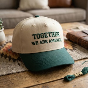 May include: A beige and green baseball cap with the text "TOGETHER WE ARE AMERICA" embroidered in green. The cap is on a wooden table with a patterned rug, a succulent plant, and a leather-bound notebook.